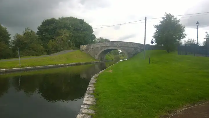 Een gebogen stenen brug voert een smalle weg over het Grand Canal bij Shannon Harbour, met op de voorgrond een grasoever met stenen rand.