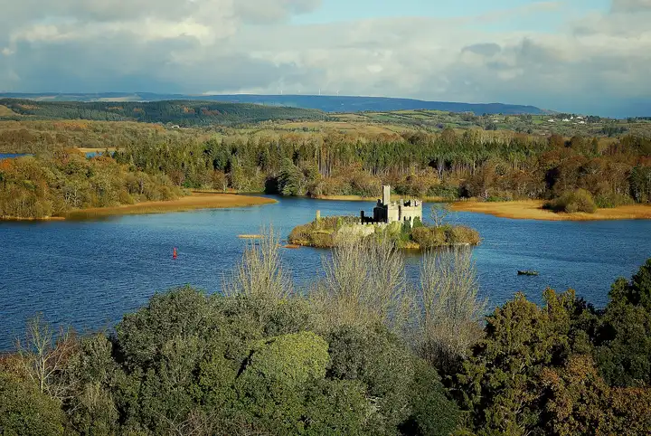 Castle Island and its castle ruin sit in a blue bay of Lough Key, seen from high above with autumn woods and distant hills behind.