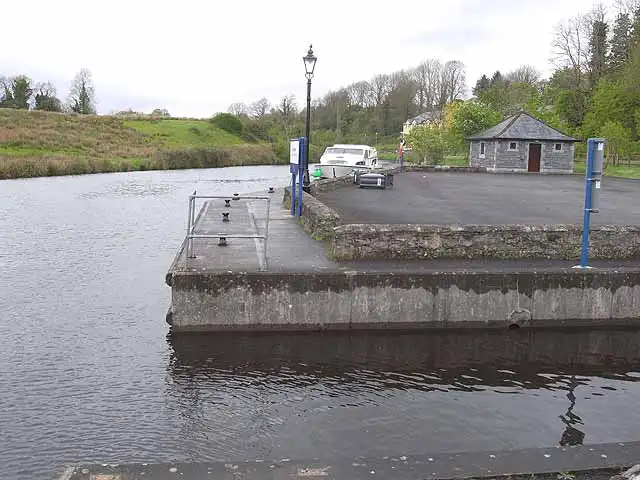 De stenen kade van de ligplaatsen in Ballinamore, met blauwe servicezuilen, een lantaarnpaal, een aangemeerde kruiser en een klein stenen dienstgebouw met schilddak.