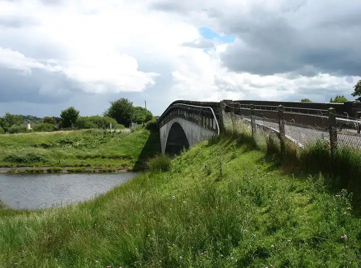 Eine Straßenbrücke mit Betonbogen überspannt in Clonlara das Wasser zwischen grasbewachsenen Ufern unter bewölktem Himmel.