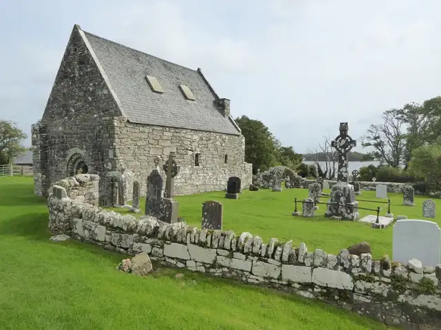 Die St.-Caimin’s-Kirche mit ihrem Friedhof auf Inis Cealtra, alte Grabsteine und ein hohes keltisches Kreuz hinter einer niedrigen Trockenmauer.