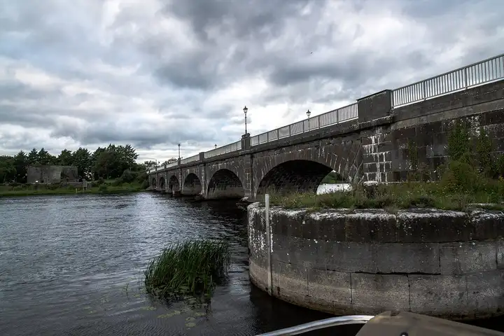 Looking along Banagher Bridge from the water’s edge, with a round stone pier standing in the river beside its arches.