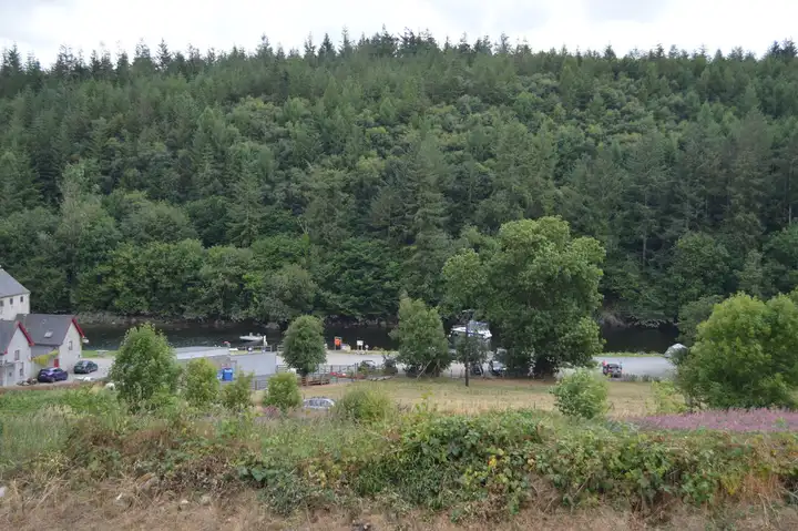 St Mullins quay and car park seen from the hill above, with boats on the tidal Barrow and steep forest on the far bank.