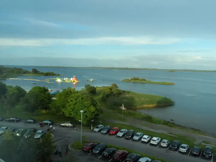 Hodson Bay seen from above on a summer evening, with an inflatable water park on the lake, a reed-fringed point, small islands and a car park along the shore.