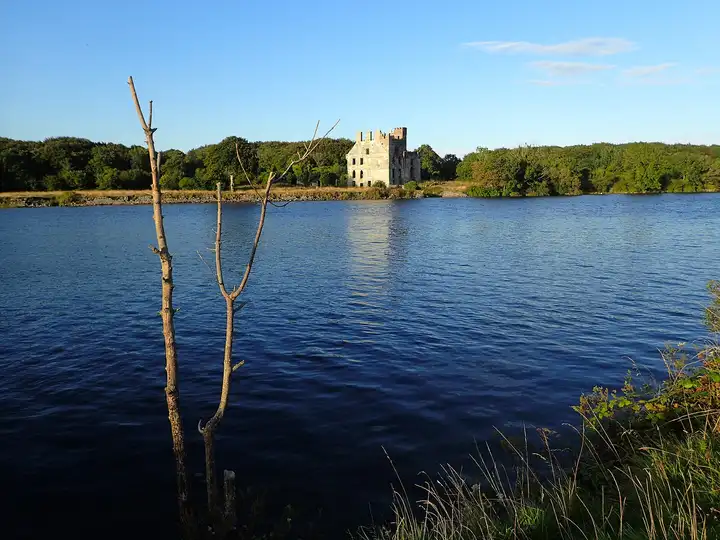 The ruin of Menlo Castle on the wooded far bank of the broad River Corrib, lit by low sunshine under a clear blue sky.