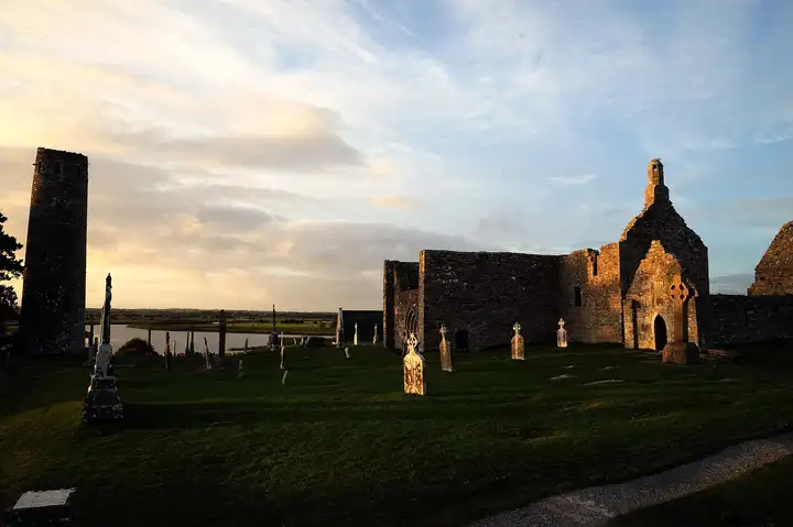 Low evening sun on the ruined churches, a round tower and grave slabs at Clonmacnoise, with the Shannon visible behind.