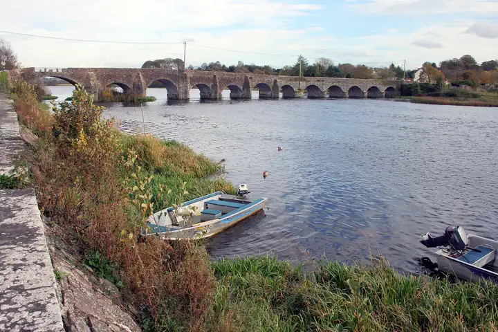 Two small outboard boats lie at a grassy bank of the Shannon, with the many-arched stone bridge of O’Briensbridge behind them.