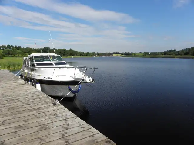 A white motor cruiser tied up at a wooden jetty at Bellanaleck, looking out over the calm river under a blue sky.