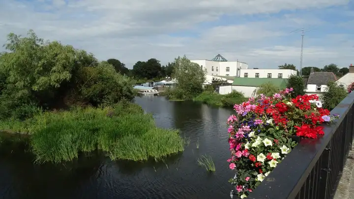 Blumenkästen an der Brückenbrüstung in Leighlinbridge über dem River Barrow, weiter hinten liegen Boote vor weißen Gebäuden am Ufer.