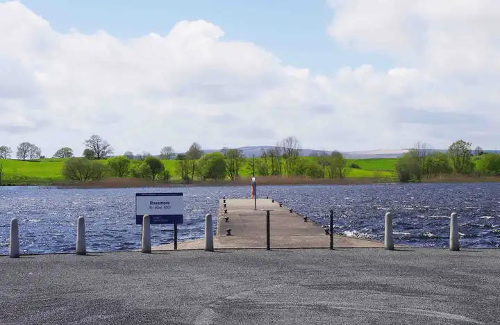 The narrow pier at Rossmore Quay running out into the lake between bollards and a name sign, with green fields on the far shore.
