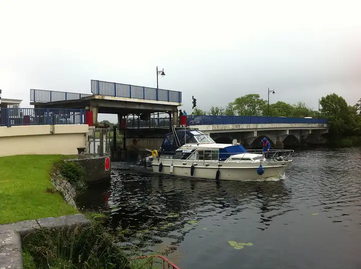 Ein Motorboot fährt an einem grauen, nassen Tag unter dem hochgezogenen Hubteil der Brücke der N5 bei Tarmonbarry hindurch.