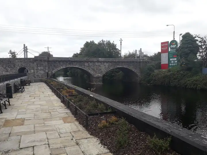 The paved riverside walk with benches and planted beds beside the Woodford River at Ballyconnell, leading to the stone arches of the town bridge.