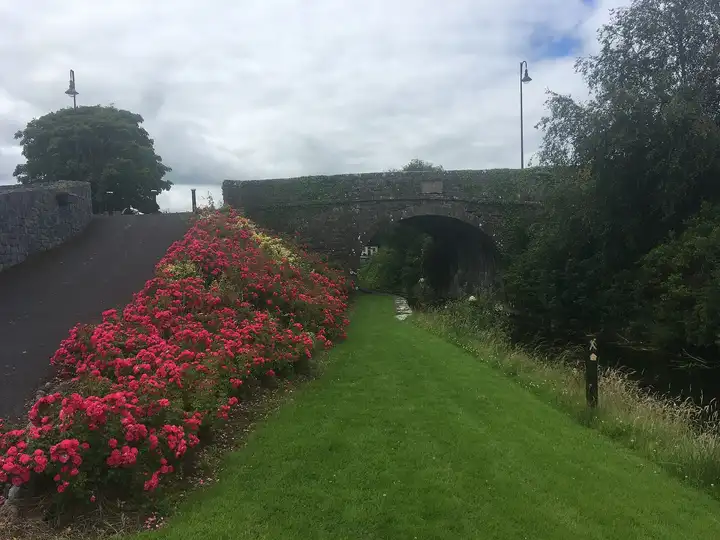 Beete mit roten Blumen säumen das Grasufer, das an einem bewölkten Sommertag zum Steinbogen der Webb's Bridge in Abbeyshrule führt.