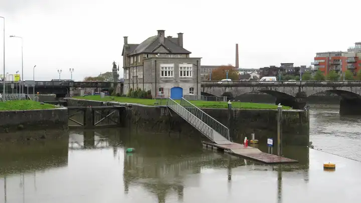 Sarsfield Lock’s gates sit in the stone quay wall beside an old house, with a gangway down to a pontoon and Sarsfield Bridge on the right.