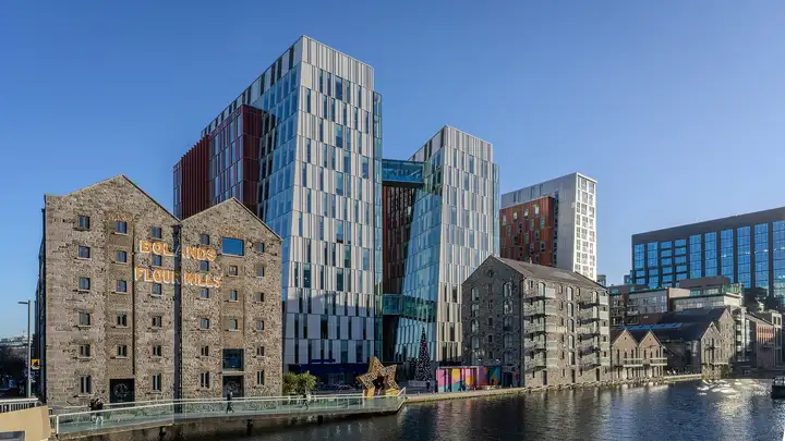 Les anciens entrepôts en calcaire des Bolands Flour Mills et de nouvelles tours de verre sur le quai du Grand Canal Dock, avec une passerelle au premier plan, sous un ciel bleu limpide.