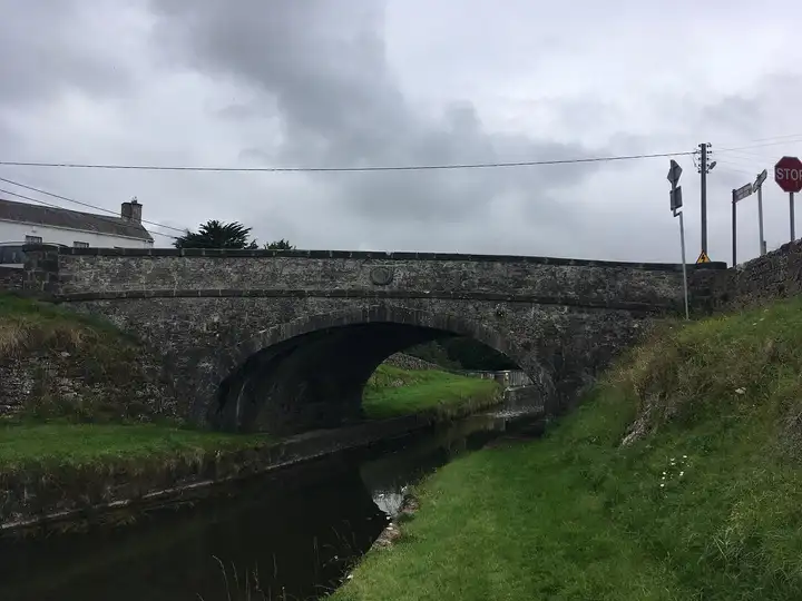 Die Molesworth Bridge in Daingean, ein breiter Steinbogen über dem Kanal, darüber Verkehrsschilder, darunter grasbewachsene Ufer.
