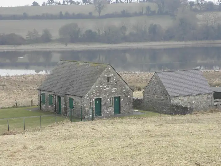 Deux petits bâtiments de pierre aux portes vertes, le centre d’accueil de l’île de Devenish, dominent le lac immobile et les champs de la rive opposée.