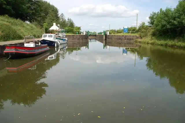 Boats moored along the bank in the calm cut leading to Portna Lock, with the lock gates and walkway at its end and a blue sign beside them.