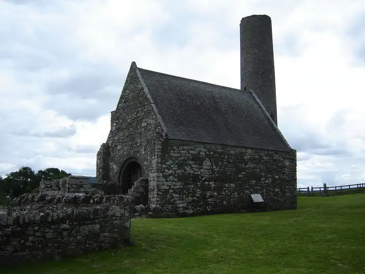 St Caimin’s Church on Inis Cealtra, a roofed stone church with a round-arched doorway, with the island’s round tower rising behind it under a cloudy sky.