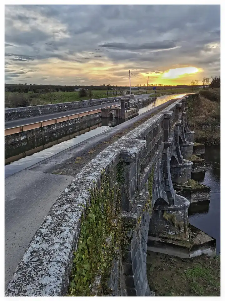 Bei Sonnenuntergang überquert der Kanal auf dem steinernen Bogen-Aquädukt von Whitworth bei Abbeyshrule den Fluss Inny, sein Wasser glänzt neben einer Straße und offenen Feldern.