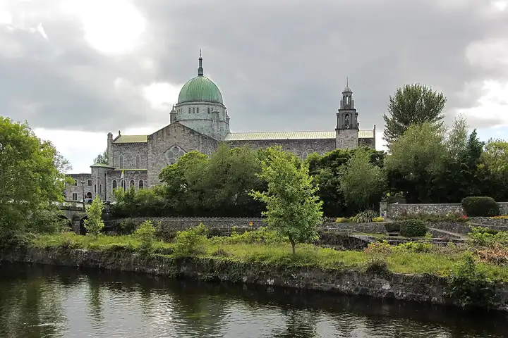 The River Corrib in Galway flowing past a grassy stone embankment, with Salmon Weir Bridge at the left edge and the green dome of Galway Cathedral above the trees.