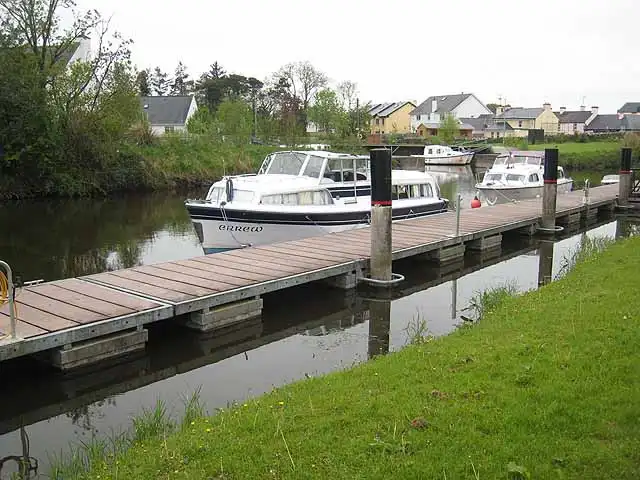 Kajütboote liegen an einem Holzsteg am Shannon-Erne-Wasserweg in Leitrim, vorn ein grasbewachsenes Ufer, dahinter Häuser.