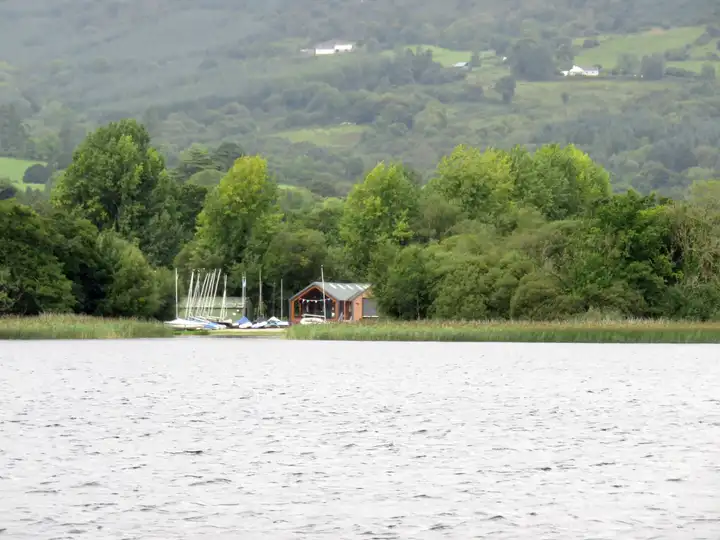 A timber boathouse and a row of dinghies on the wooded Clare shore near Ballycuggaran, seen across the lake.