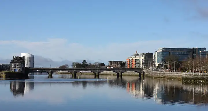 Sarsfield Bridge and the modern buildings on its banks are mirrored in the still Shannon under a clear blue sky.