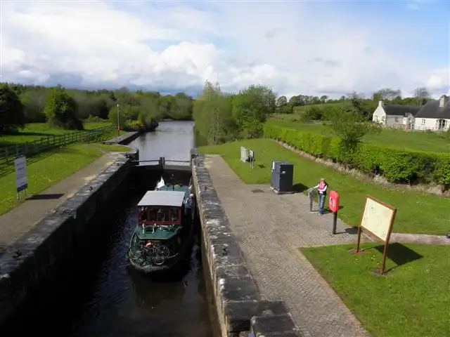 A green barge in Corraquill Lock (Lock 1) on the Woodford River, seen from above the gates, with the paved lockside, a cottage and the river stretching away.