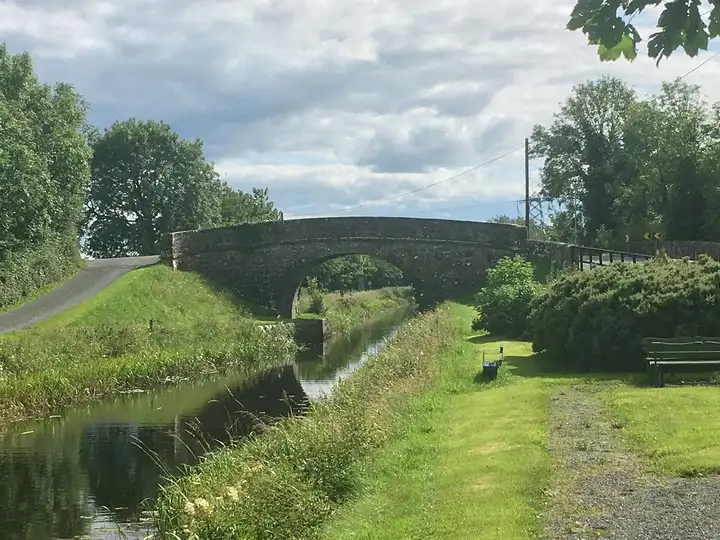Der Kanal fließt unter dem Steinbogen der Scally's Bridge bei Abbeyshrule hindurch, rechts ein gemähter Treidelpfad, eine Bank und ein Picknicktisch.