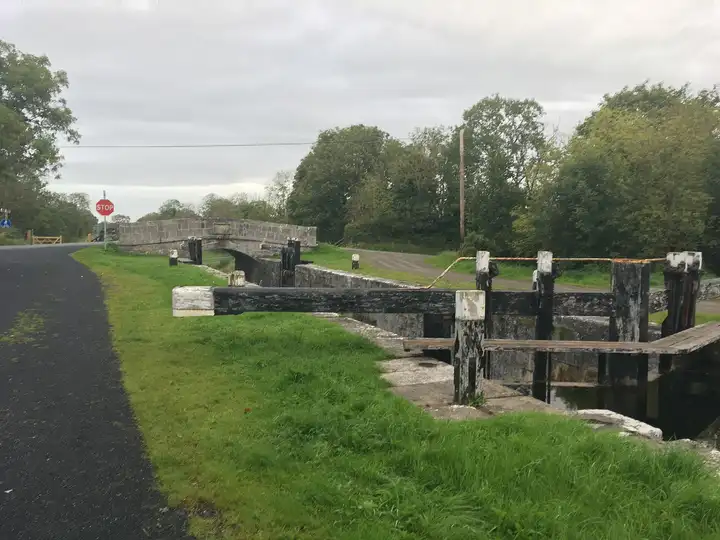 De zwarte houten balansbalken van sluis 44 bij Killashee liggen naast een grazig jaagpad, met daarachter de stenen boog van Savage Bridge onder een grijze lucht.