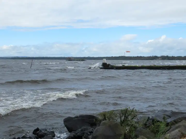 Wellen rollen am Lough Neagh bei Antrim heran und brechen sich an einem niedrigen steinernen Wellenbrecher mit Windsack, unter blauem Himmel mit weißen Wolken.