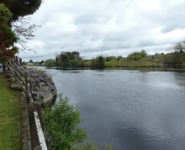 A timber rail and a rock-armoured bank follow the broad River Erne at Belleek, with houses in the distance and green banks opposite under a cloudy sky.