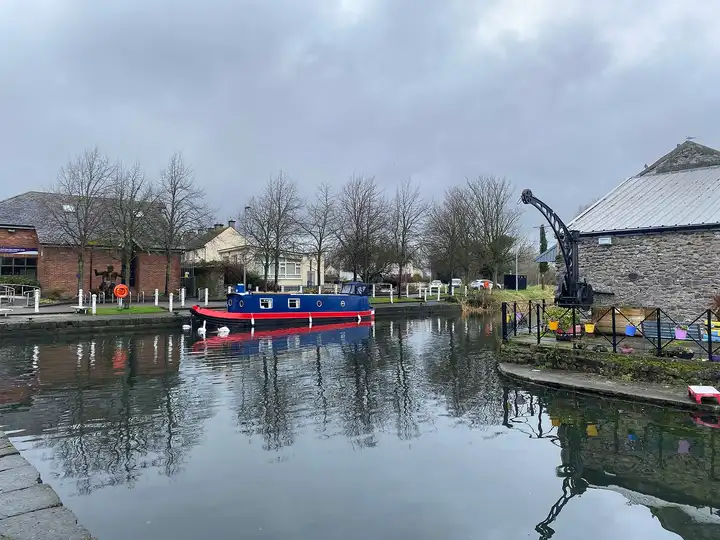 Naas Harbour on a grey day, with a blue-and-red narrowboat and swans by the quay, an old harbour crane and the stone canal stores on the right.