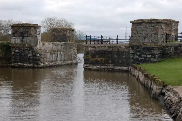 The square stone piers of the former railway bridge at Toome rise from the water on both sides of a narrow channel, with railings and a grassy bank beside them.