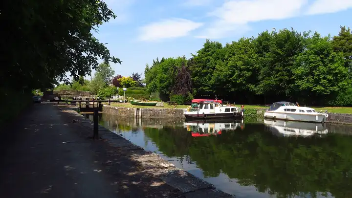 Two cruisers lie moored in Coolnahay Harbour, mirrored in still water, with a green rowing boat on the lawn, Lock 26 and its bridge on the left and tall trees behind.