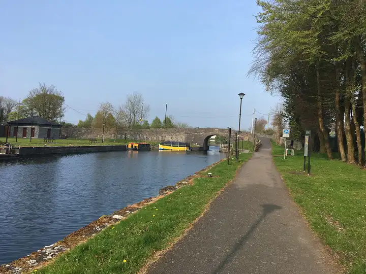 A paved towpath runs beside the canal to the stone arch of Scanlan's Bridge, where two colourful boats are moored by the harbour lawn and a stone service building.