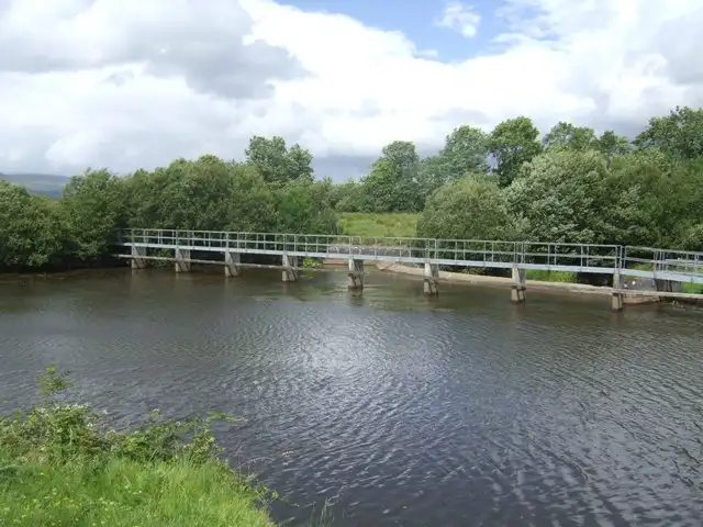 A long footbridge with metal railings crossing the overflow weir near Castlefore Lock, with trees and summer clouds behind.