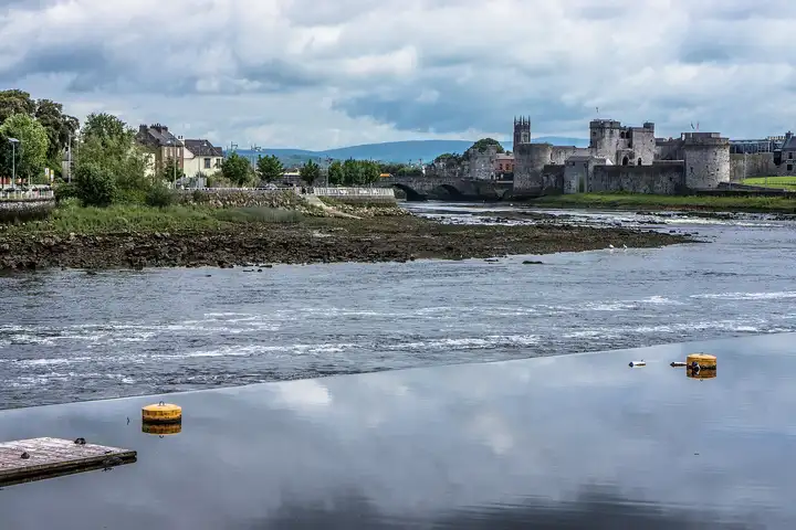 The Shannon tumbles over the Curragower Falls past King John’s Castle and Thomond Bridge, with yellow buoys on the calm water in front.