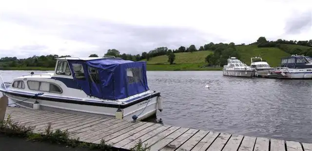 A motor cruiser with a blue canopy moored at a wooden jetty at Bellanaleck, with more cruisers moored further along the river.