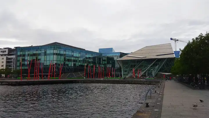 Le plan d’eau du bassin devant Grand Canal Square, avec ses mâts lumineux rouges inclinés, ses immeubles de bureaux vitrés et le Bord Gáis Energy Theatre aux formes anguleuses, sous un ciel gris.