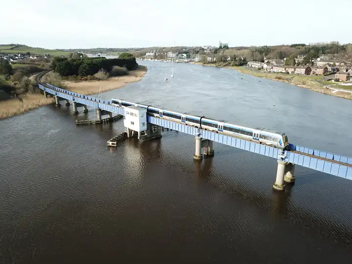 Vue aérienne d’un train de voyageurs franchissant le long pont ferroviaire bleu sur la Bann à Coleraine, avec la rivière et des bateaux au mouillage vers l’aval.