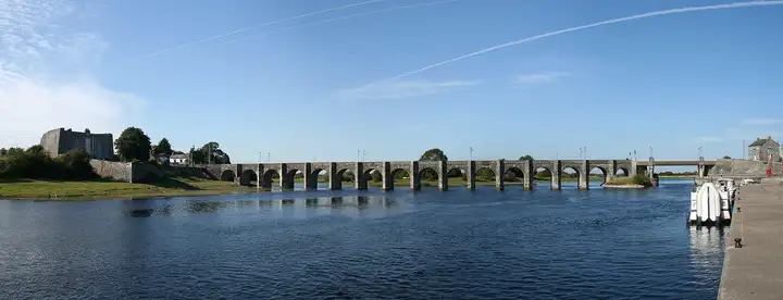 Vue large du long pont aux nombreuses arches de Shannonbridge sous un ciel bleu, du fort sur la rive du Roscommon jusqu’à un bateau amarré au quai à droite.