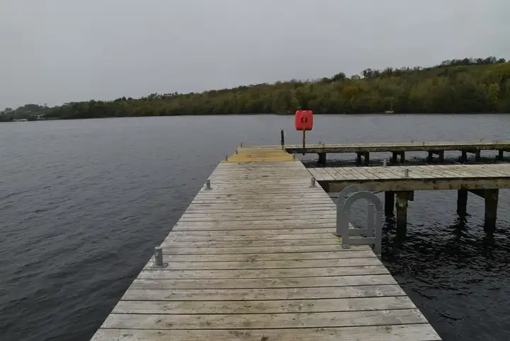 A timber jetty with a lifebuoy runs out into the lough from the Castle Archdale forest shore, facing a wooded far bank under an overcast sky.