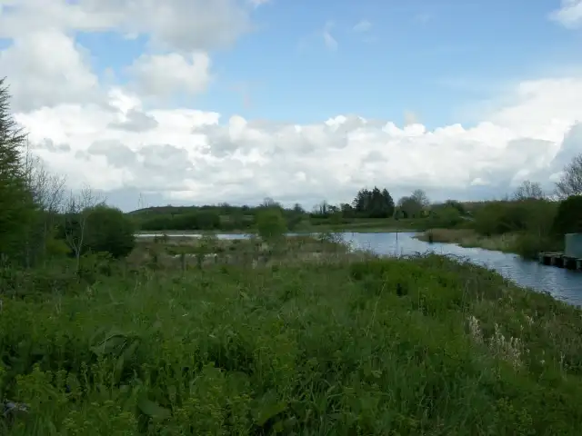 De Shannon–Erne Waterway kronkelt door biezen en weiland bij de sluizen van Kilclare, met rechts het uiteinde van een steiger onder een wijde wolkenlucht.