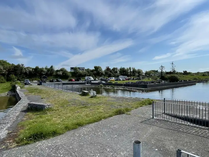 Annaghdown Pier par une journée ensoleillée, avec des garde-corps le long de la cale de mise à l’eau, le bassin abrité et des voitures garées sur la rive.
