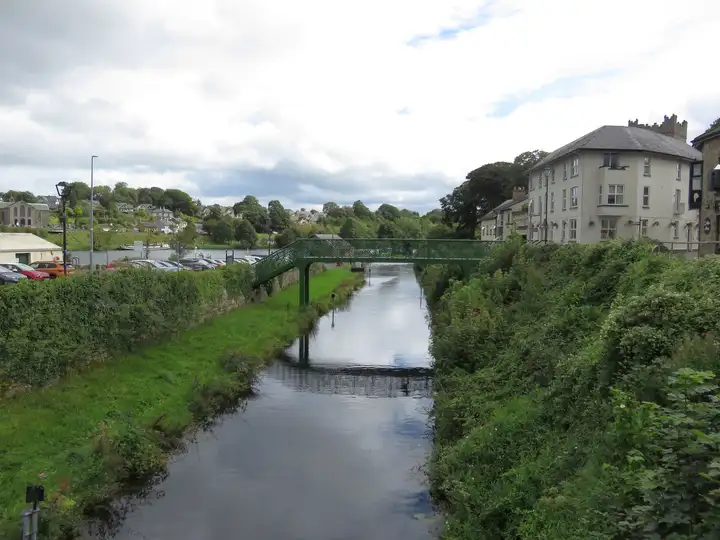 Le court canal de Killaloe entre des berges herbeuses, enjambé par une passerelle verte en treillis, avec le Shannon et Ballina en arrière-plan.