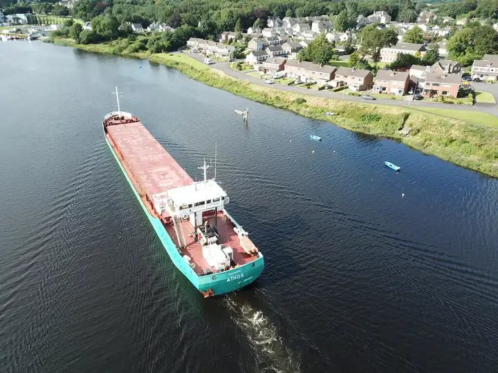Vue aérienne du cargo Athos sur la Bann soumise à la marée, juste en aval du pont ferroviaire de Coleraine, le long d’une rive bordée de maisons, avec de petits bateaux au mouillage.