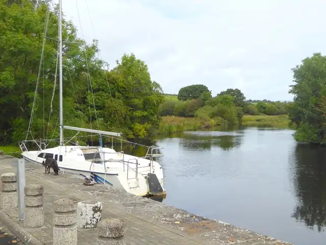A small sailing yacht moored at the stone quay of Tuamgraney on the tree-lined Scarriff River, with a dog on the quayside.