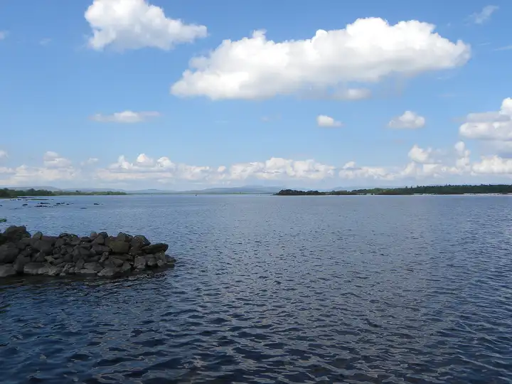 The wide open water of Lough Corrib at Knockferry under a blue sky with white clouds, with a rocky spit in the foreground and low wooded shores far off.